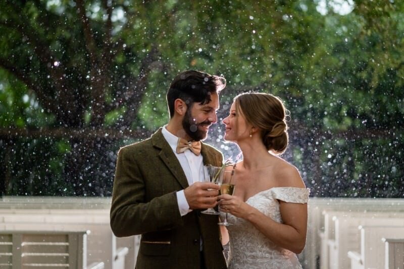A newlywed couple shares a moment toasting with champagne in a beautiful rainy outdoor setting