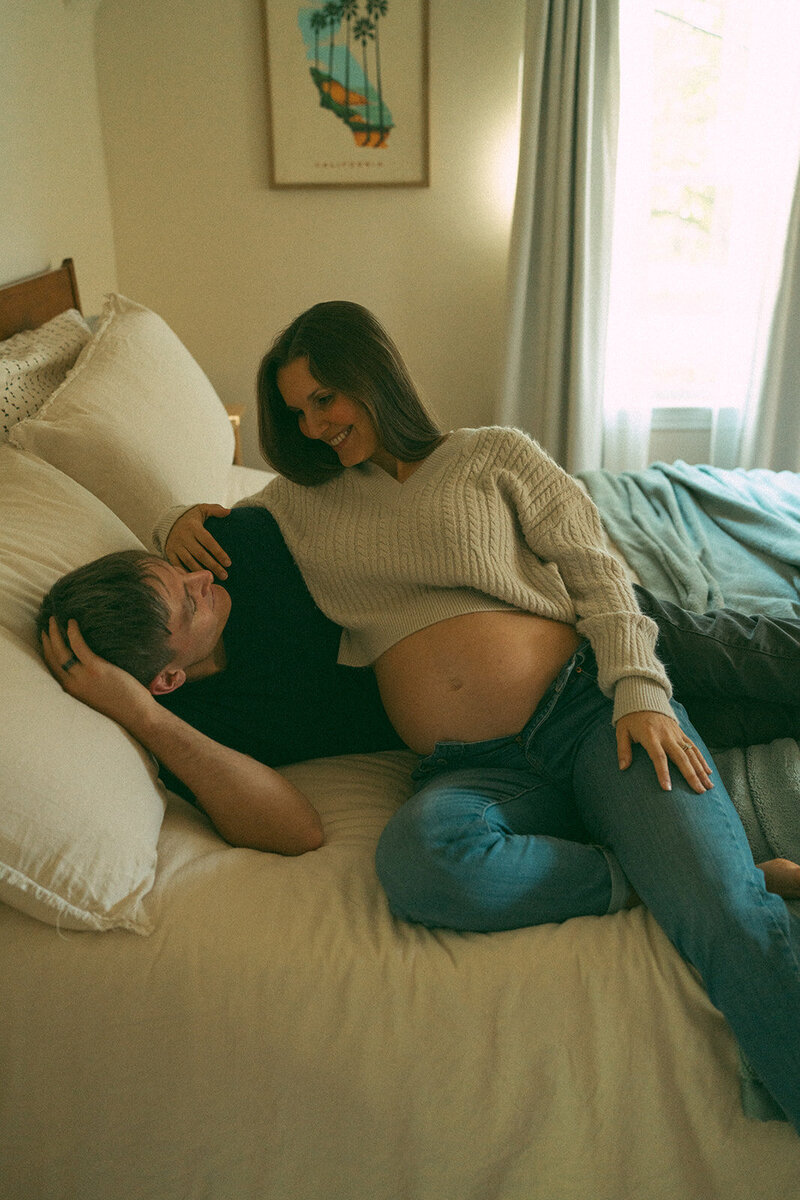 Pregnant woman and her partner cuddling together on a bed during a relaxed, documentary-style maternity photo session

