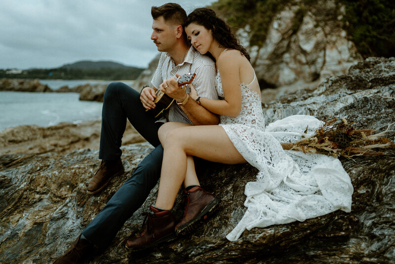 N & M sitting side by side on a large rock on the beach, N holding onto M's chest while M looks out onto the water while playing the ukelele