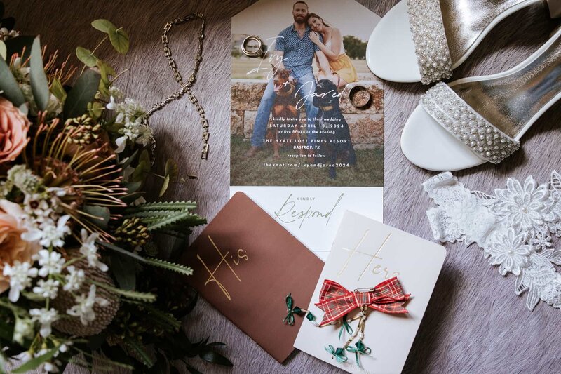 A flat lay photo featuring a rustic wedding invitation, His and Hers vow books with festive accents, bridal shoes with pearl embellishments, a lace garter, jewelry, and a unique floral arrangement, styled for a warm and intimate wedding celebration.