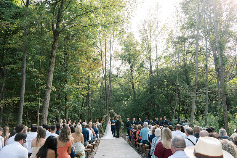 Forest wedding ceremony with a hexagon arbor, wooden benches, and guests gathered under tall trees at Bear Ridge Destination.

