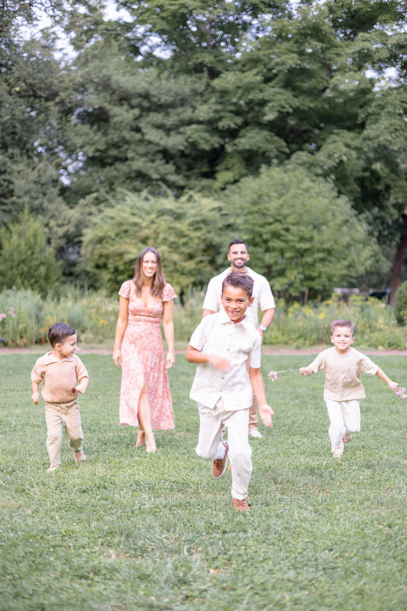A family of five enjoying time together in a lush park, with three young boys running toward the camera and their parents smiling behind them under soft, golden light.