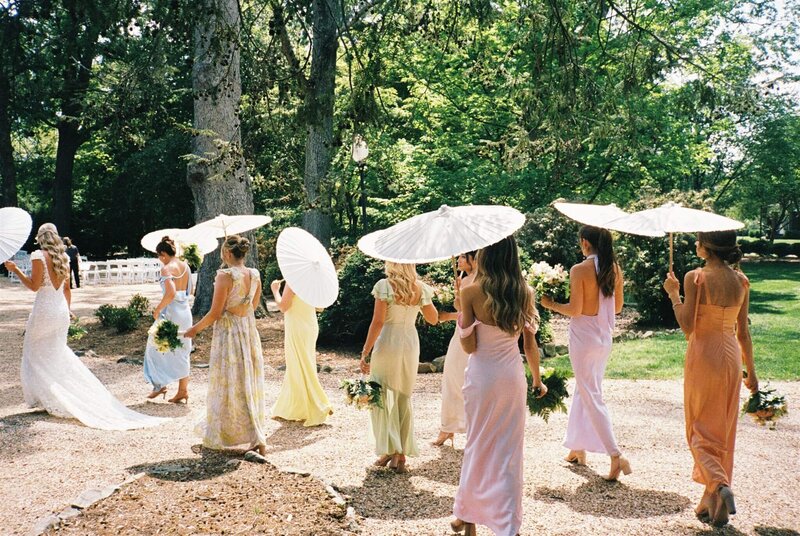 A group of bridesmaids in colorful dresses walk in lines behind the bride holding white umbrellas