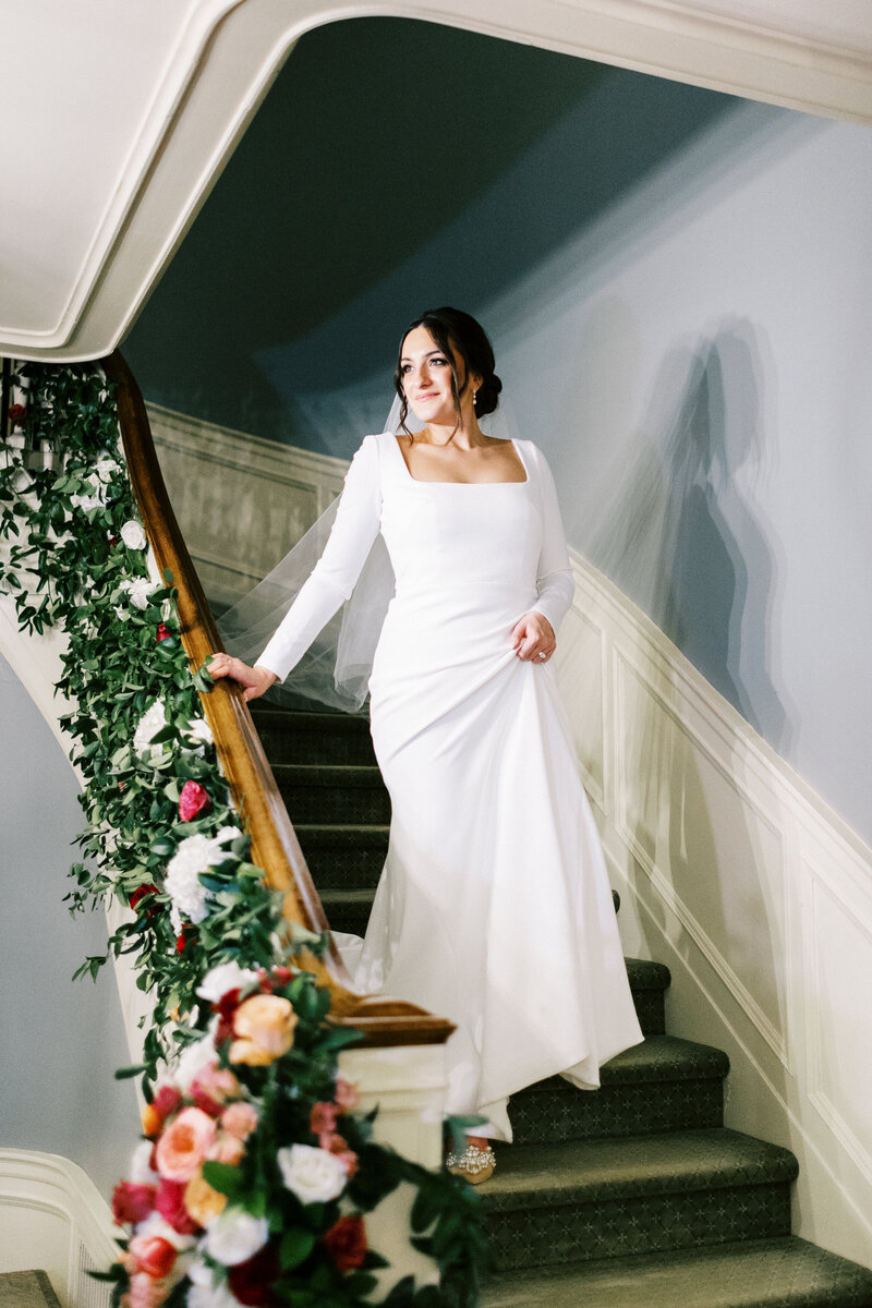 Bride with a smile walking down the decorated stairs with flowers in her elegant wedding dress at St. Paul College Club, captured by Minnesota wedding photographer.