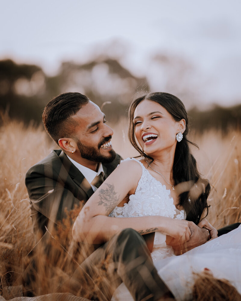 Bride & groom sitting amongst long grass laughing as they cuddle