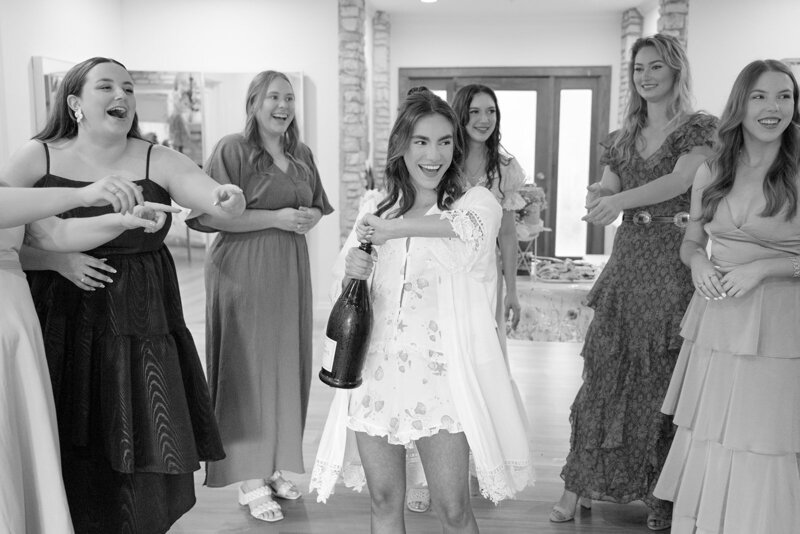 Candid bridal prep photo of bride holding champagne bottle with bridal party reacting taken by Austin photographer , Erin Marcin