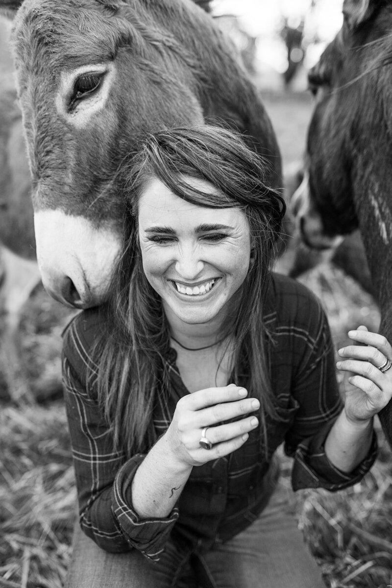 Woman laughing with a donkey nuzzling her shoulder, representing the work of lifestyle photographer Heather of Among the Oaks. 