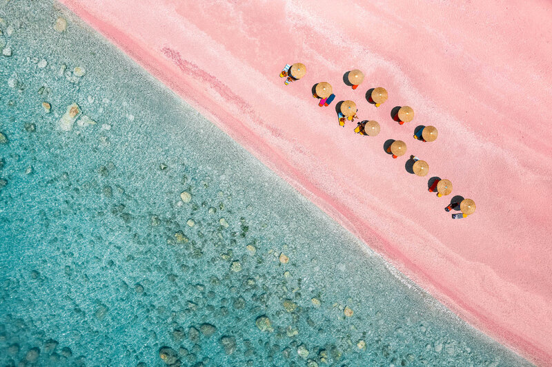 Aerial view of beach gazebos along a pink sand beach beside clear turquoise water.