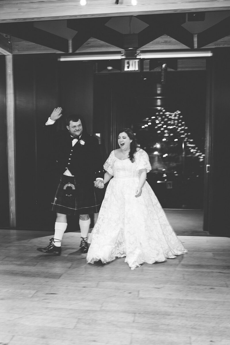 Black and white image of Scottish groom and Jewish bride walking into their wedding reception while holding hands raised and cheering  in the ballroom at District Winery in DC 