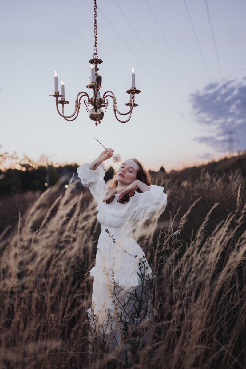 Woman wearing white gown poses in a grassy field with elegant chandelier hanging above her.