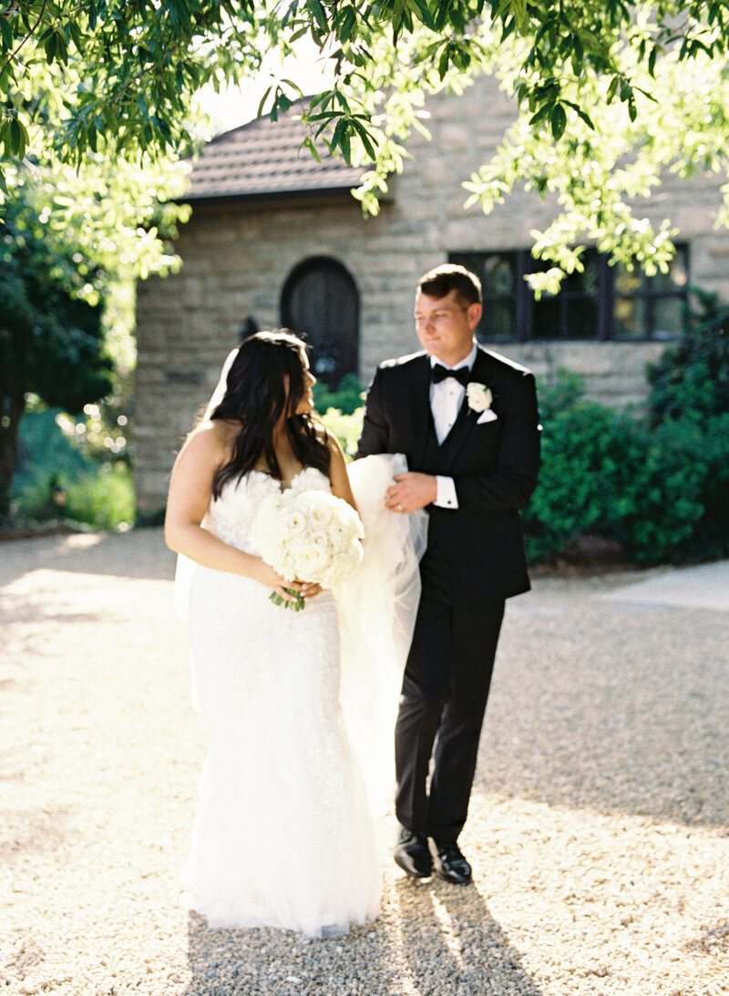 Out of focus image of a bride and groom walking together while groom holds the brides dress and smiles at his bride.