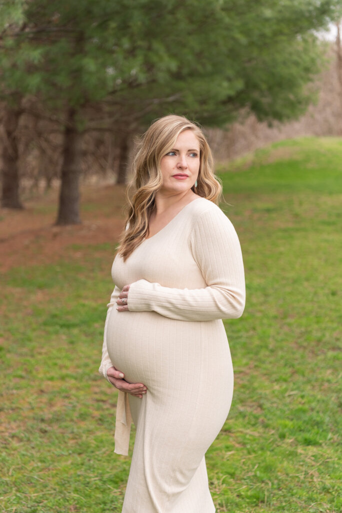 A pregnant women wearing a beige colored dress holding her belly at a park.