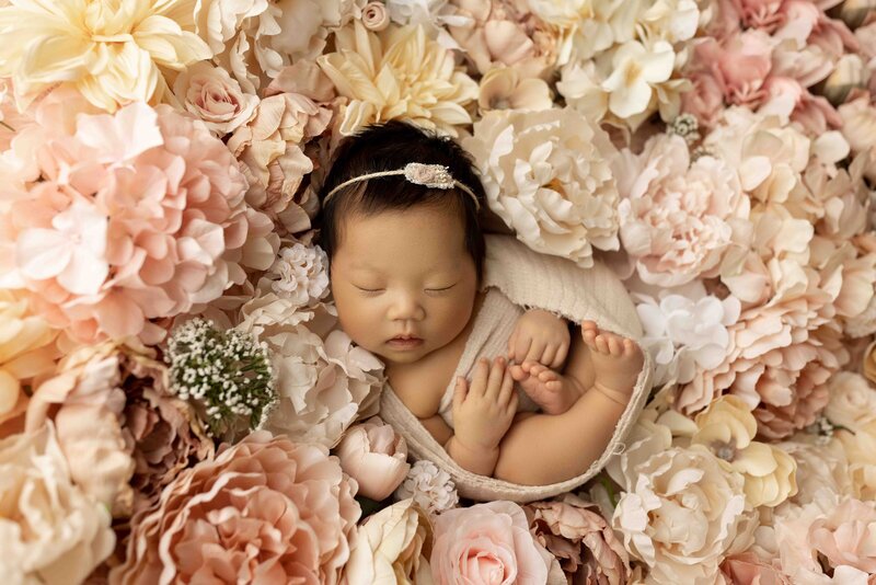 Newborn baby girl from London Ontario curled up for newborn photoshoot. She is sleeping on a bed of blush and cream flowers