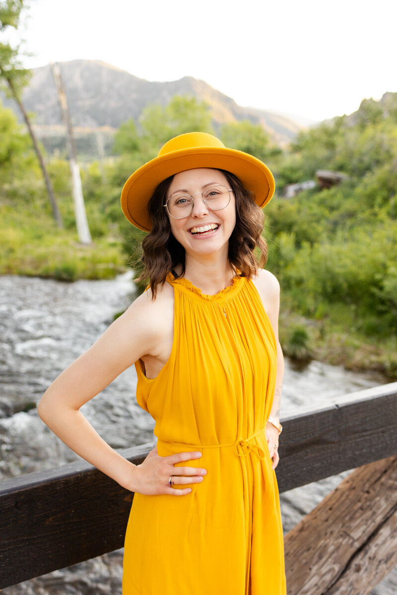 A woman in a yellow dress and yellow hat stands with her hands on her waist and smiles at the camera.