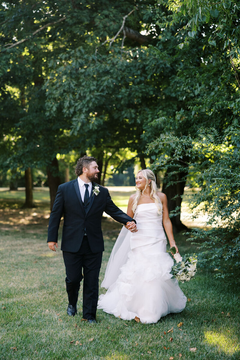 Kaiela and Zach hold hands on the lawn of Boxwood Estate. Kailea is wearing a white gown with her hair down, holding a white bouquet. Zach is wearing a classic black tuxedo with a white rose boutonnière on his left side.  
