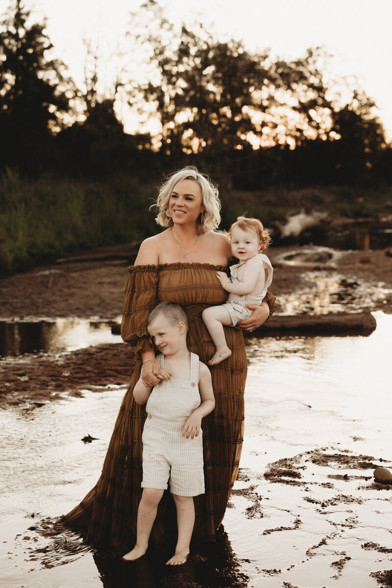 Kim stands in a long dress stands outdoors, holding a toddler while another young child stands beside her. The background features a creek area with trees, capturing the essence of Wollongong motherhood sessions.