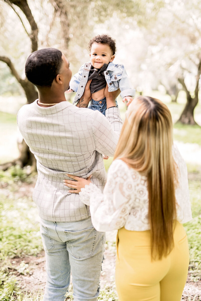 A family with their backs turned to the camera in a sunny lush pathway. The dad holds up his baby who smiles at the camera.