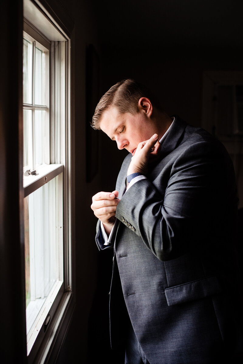 Groom standing at a window putting his cufflinks in