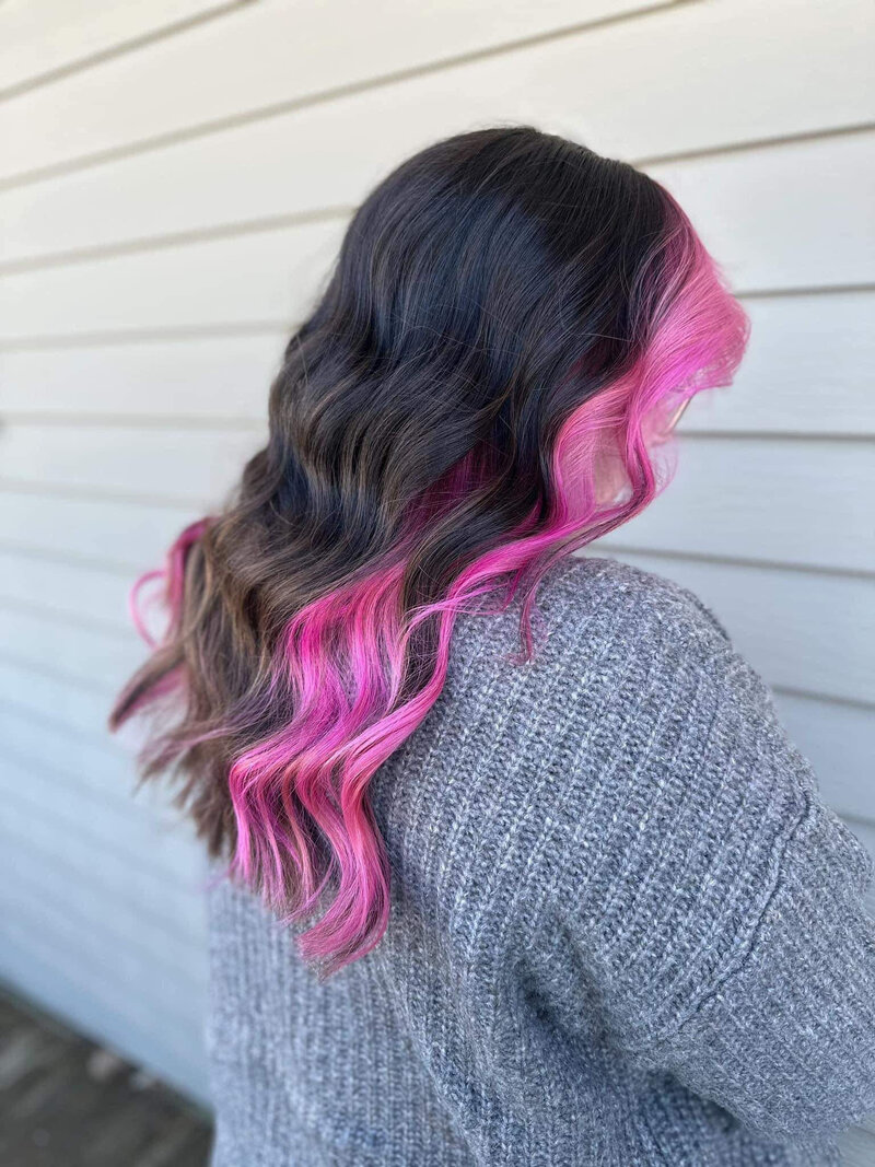 A brunette with a pink color block in the front of her hair, standing outside of a tan building