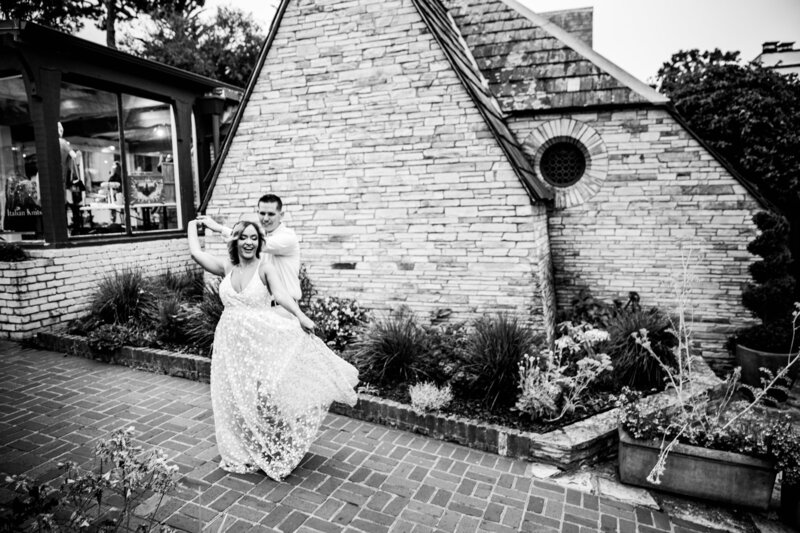 A joyful couple dances together in a charming brick courtyard, the bride twirling in her flowing gown while the groom playfully embraces her from behind, set against a rustic brick building