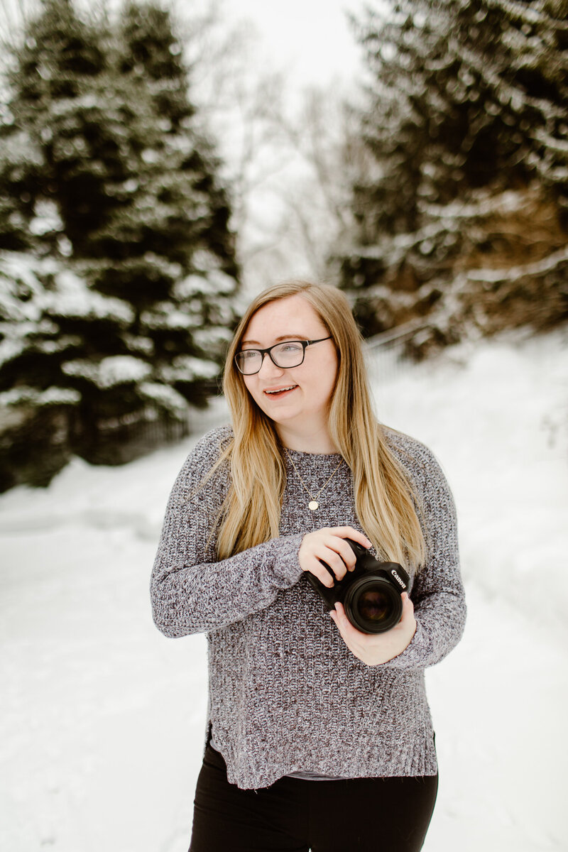 Blonde woman holds camera while smiling surrounded by snow.