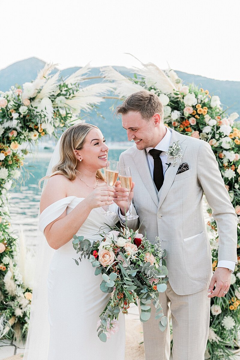 Bride and groom share a celebratory champagne toast in front of a pampas grass floral arch by the water, smiling joyfully after their ceremony