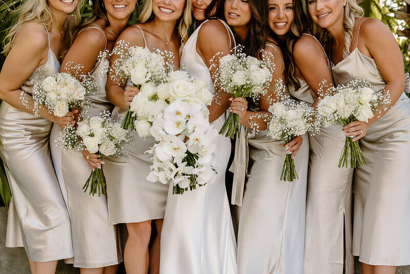 Bride and bridesmaids in neutral satin dresses pose with white rose bouquets in front of a tall cactus at Acre Resort, Cabo wedding.