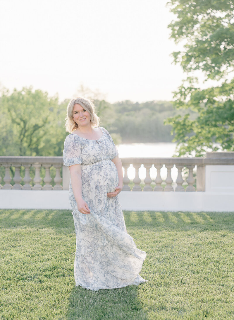 A pregnant mom with blonde hair wearing a blue floral dress holds her bump and smiles at Indianapolis Maternity Photographer Brittney Lear.