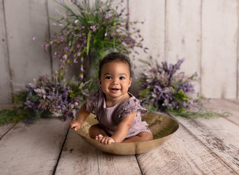 baby girl smiling sitting in a gold hammered bowl with purple florals for milestone photo session