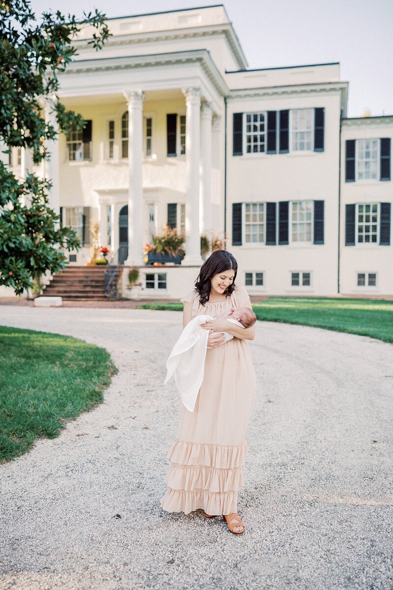 Mother with tan flowing dress and brown hair stands in front of Oatlands Historic Home holding her newborn baby swaddled in white, photographed by Northern VA Newborn Photographer Marie.
