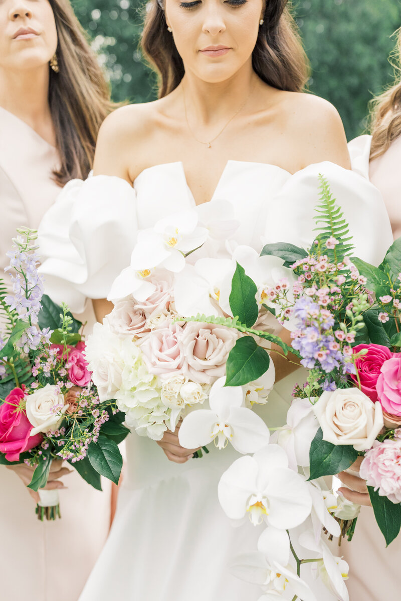 Bride in off-the-shoulder gown holding a bouquet of blush roses, white orchids, and greenery with pops of pink