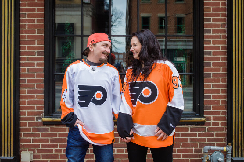 Engaged couple dressed in Flyers attire laughing together, sharing playful and candid moments during their sports-themed engagement session.