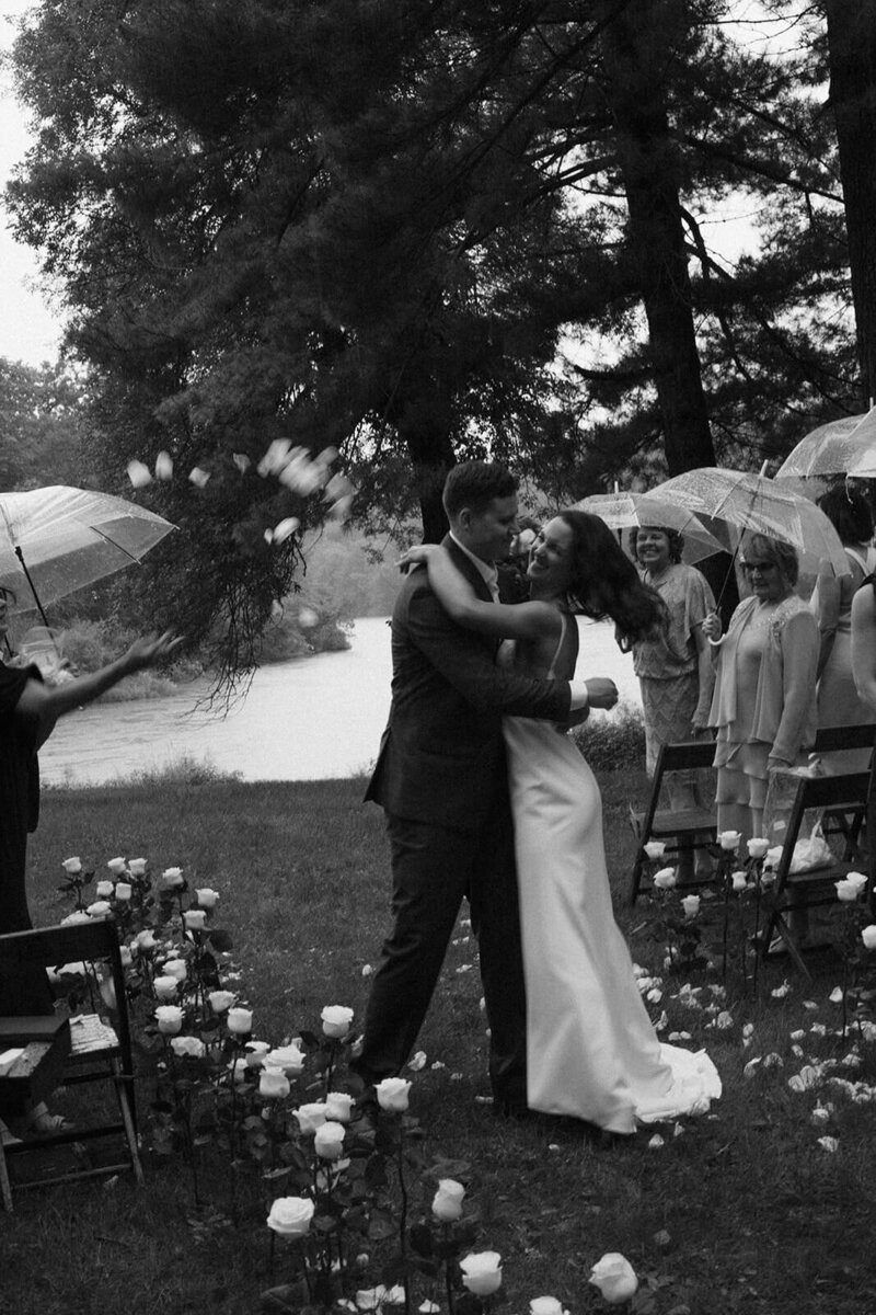 Bride and groom twirl in the aisle as wedding guests look on with umbrellas and throw flower petals in the air