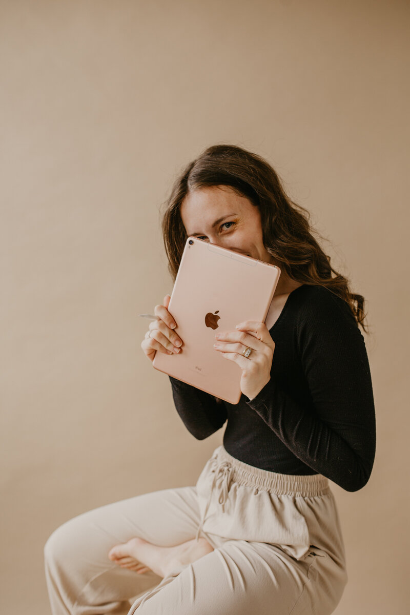 A confident coach and UGC creator peeking behind her iPad, sitting on a stool with comfy looking linen pants and a sleek black long sleeve. 