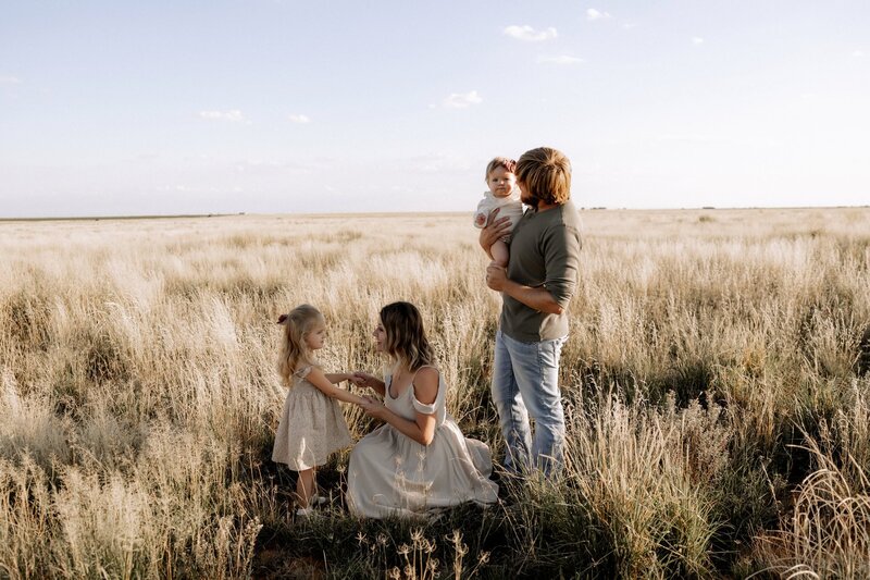 Family of four in a golden field during sunset—mother kneeling with daughter, father holding baby, captured in a candid, natural moment.