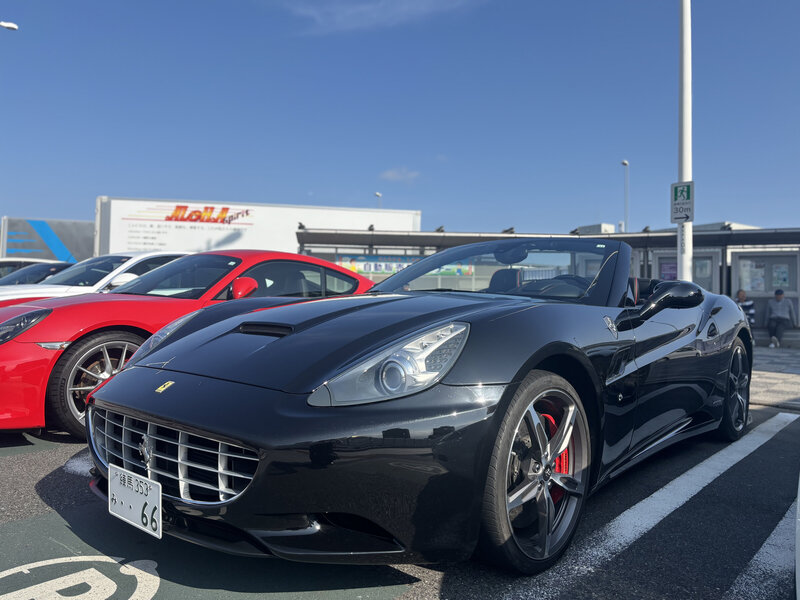 Front-side view of a black Ferrari California parked in Tatsumi Parking Area under a clear blue sky, with other sports cars in the background.