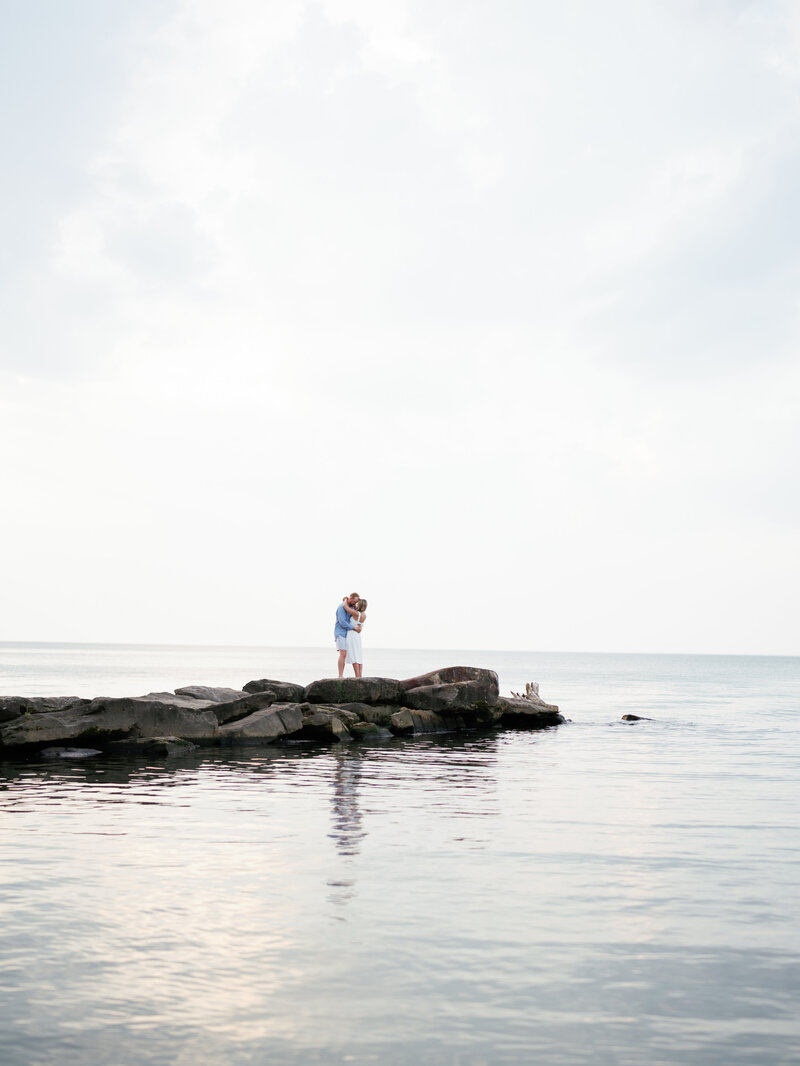 Couple stands on rocks surrounded by Lake Erie at Edgewater Beach engagement session

