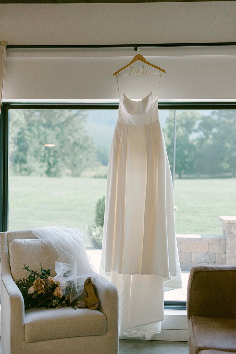 Gorgeous bridal gown hanging in a large window with hints of light creating a soft glow around the dress.