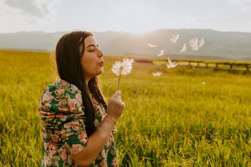 Senior, Madeline blows on a dandelion stem with a brightly lit field in the background.
