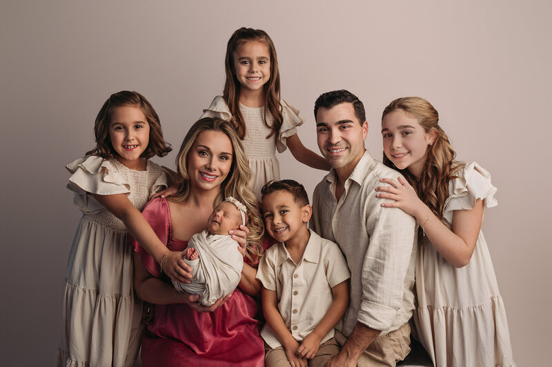 beautiful family of seven posing with a newborn baby girl in front of a cream backdrop