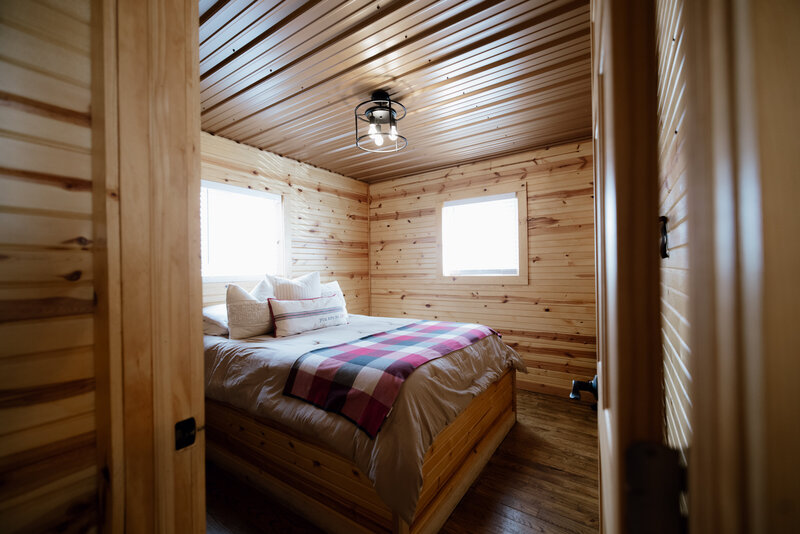 Cozy wooden cabin bedroom with natural pine walls, a plaid blanket on a queen bed, and soft natural light from two windows at Bear Ridge Destination.

