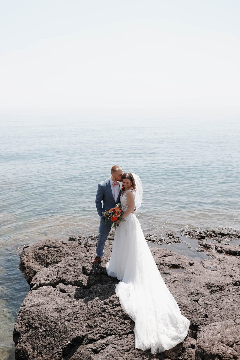 A newlywed couple standing close on a rocky shoreline 