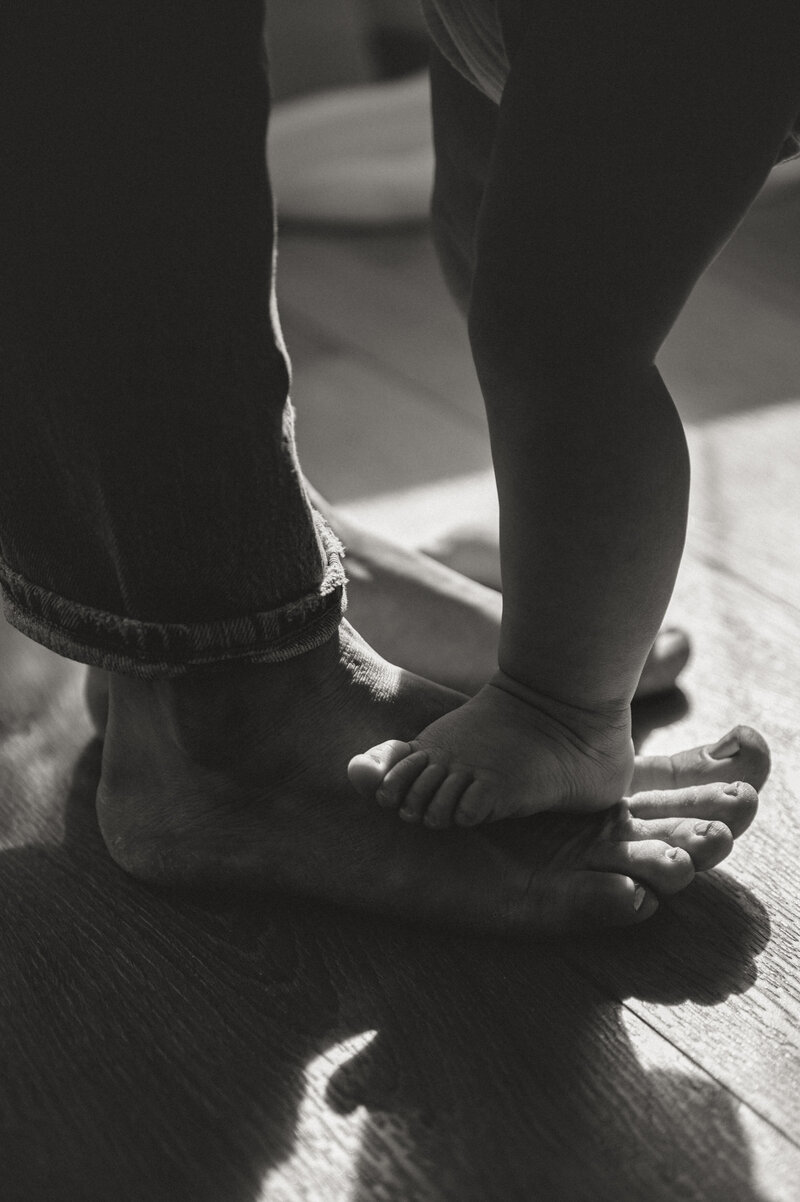 A toddler's feet standing on their grandparent's feet in a sunlit studio. 