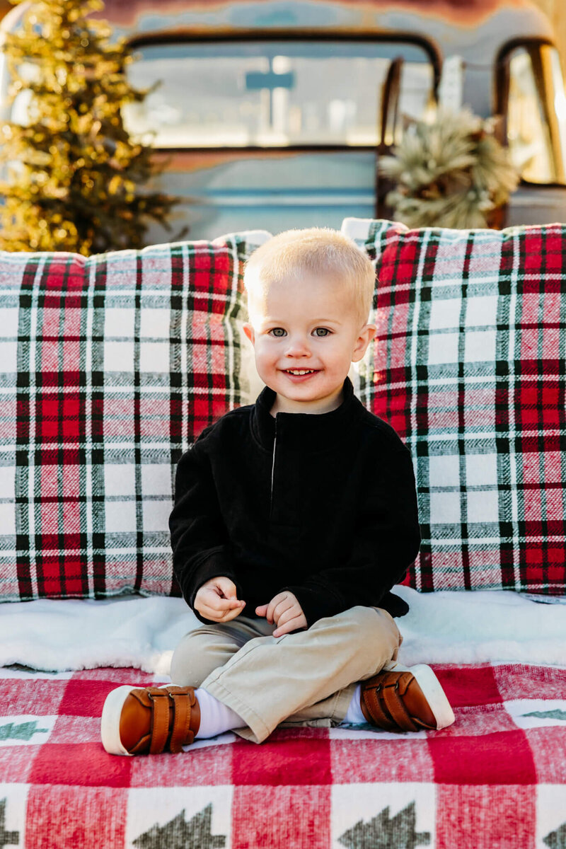 adorable little boy sitting on a plaid blanket in an old blue truck that is decorated for Christmas captured by Ashley Kalbus Photography