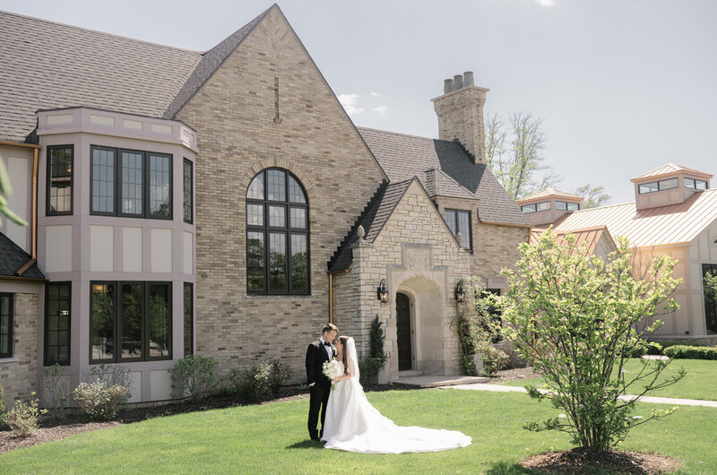 A bride with a long white veil and groom in a black tuxedo stand in the front of a stone manor on their spring wedding day in Grand Rapids.