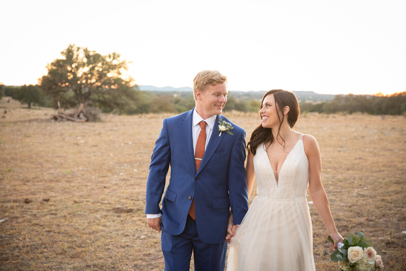 Austin wedding photographer captures a stunning moment of a bride and groom walking through a field at the enchanting sunset.