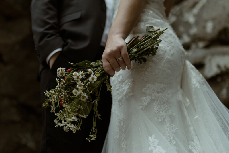 close-up image of a bride in a lace wedding gown holding a hand-picked bouquet of wildflowers, including daisies and greenery, with the stems casually gathered in her hand. The groom stands beside her in a dark suit with one hand in his pocket, partially visible in the background. The texture of the bride’s gown features delicate floral embroidery and soft tulle, which contrasts beautifully with the earthy, natural tones of the bouquet. The couple stands in front of a rocky, outdoor backdrop, capturing the organic and unpolished beauty of their nature-inspired wedding day.