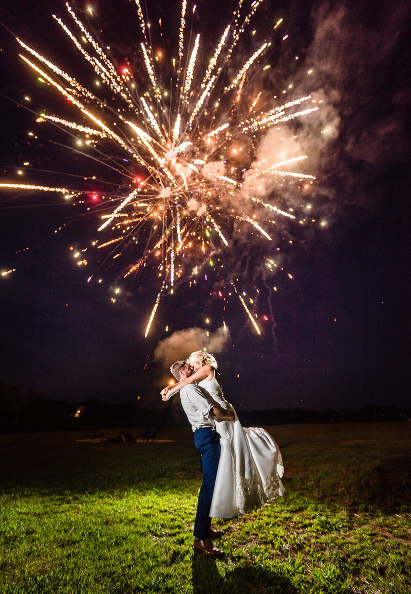 A groom lovingly lifts his bride and holds her up as fireworks explode in the background.