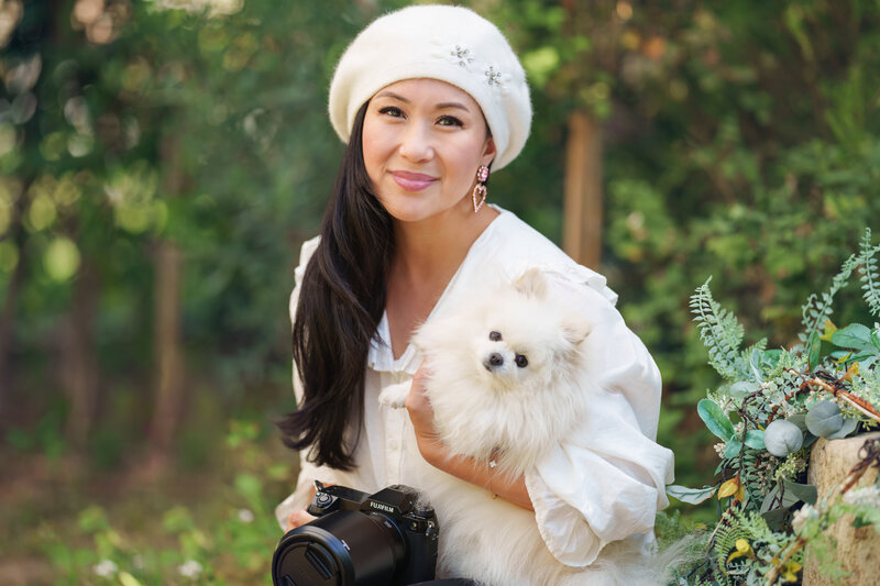 Asian woman holding a small white pomeranian dog and a black Fujifilm camera sitting in a garden while wearing a white beret, pink heart earrings, and a white button up shirt with a peter pan collar.