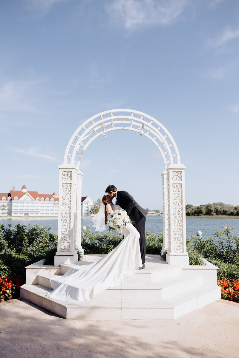 Bride and groom embrace, as bride looks softly into groom’s face as he looks down at her and brushes her hair to the side of her face. Her hair is in a soft updo with a beautiful white veil pinned behind. Groom wears a black tux and boutonniere of white roses. Photo taken by Orlando Wedding Photographer Four Loves Photos and Film.